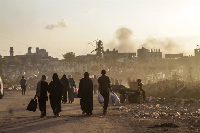 October 17, 2024, Gaza, Palestine: Palestinians walk along a street which was bombed by the Israeli army as smoke rises in Gaza City.