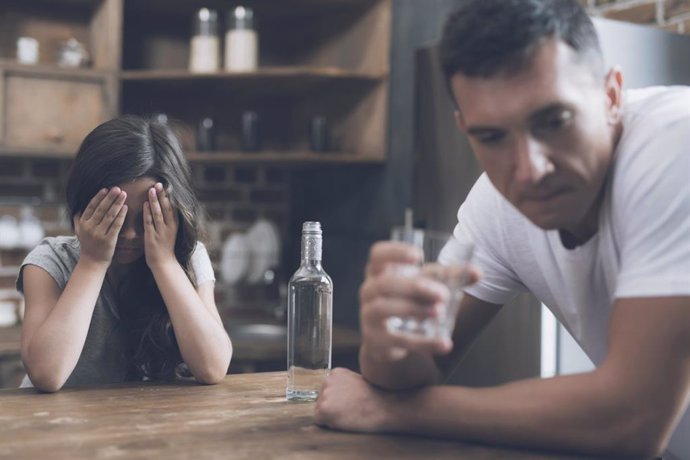 Archivo - Una niña está llorando cubriendo su rostro con sus manos, mientras que su padre bebe.