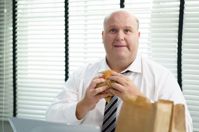 Archivo - Hombre con sobrepeso comiendo hambuerguesa en la oficina, mirando a la cámara.