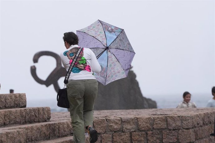 Archivo - Una mujer camina con un paraguas junto al Peine del Viento