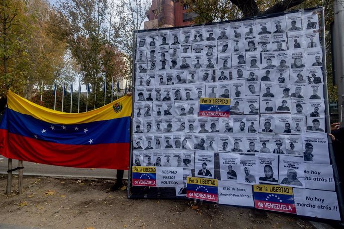 Un cartel de protesta con fotografías junto a una bandera de Venezuela durante una manifestación de venezolanos en Madrid, frente a la comisión del Parlamento Europeo, a 1 de diciembre de 2024, en Madrid (España). La protesta, convocada “por la libertad y