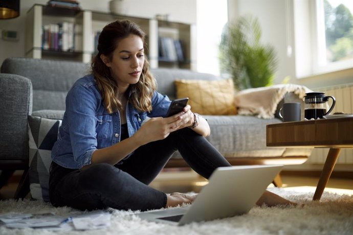 Archivo - Mujer joven que usa el teléfono móvil y el ordenador portátil en casa.