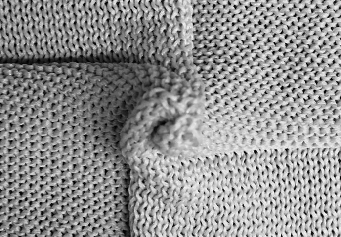 Um close-up de um tecido de malha que se dobra automaticamente, demonstrando como padrões de pontos específicos (pontos retos e reversos) codificam regras geométricas que ditam a forma tridimensional do material.