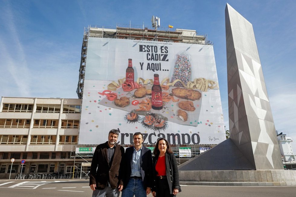 Ada Bernal, directora regional de Relaciones Institucionales de Heineken España, el alcalde de Cádiz, Bruno García, y el pintor Pepe Baena, junto a la lona desplegada por Cruzcampo con motivo del Carnaval de Cádiz