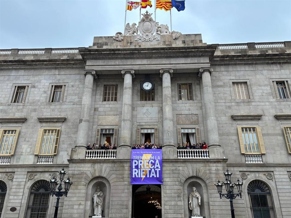 El alcalde Jaume Collboni, representantes del gobierno, de grupos políticos y de entidades han desplegado una pancarta en el balcón del Ayuntamiento con el lema de este año para el 8M 'Trenquem la precarietat'