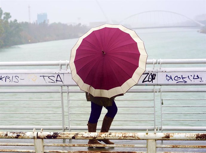 Archivo - Un persona se protege de la lluvia y el viento caminando por la pasarela  peatonal de la Cartuja en un día lluvioso y con fuertes rachas de viento en la provincia. En Sevilla (Andalucía, España), a 09 de febrero de 2021.