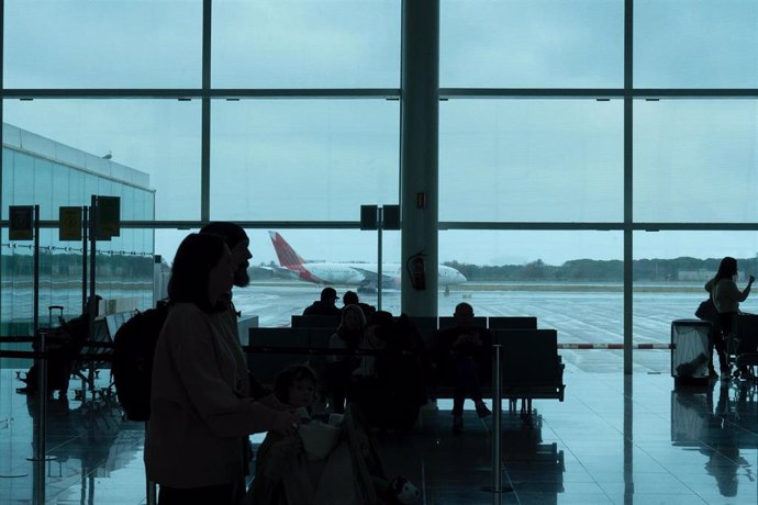 Archivo - Varias personas esperan sentadas frente a un avión aparcado en la pista de un aeropuerto.