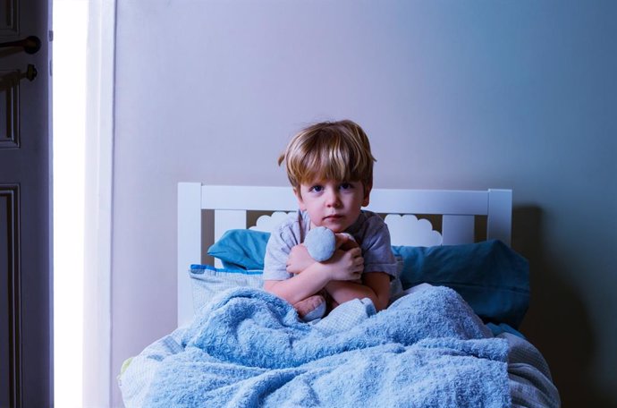 Archivo - Little blond calm boy sit in the bed holding plush toy covered in blanked near slightly open door with day light