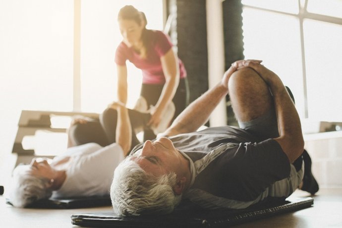 Archivo - Casal de idosos se exercita em centro de reabilitação. Personal trainer ajuda casal de idosos a fazer alongamento no chão. Foco no homem.