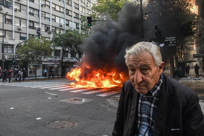 12 de março de 2025, Buenos Aires, Argentina: Um aposentado caminha pela rua enquanto, ao fundo, um carro pega fogo durante a manifestação no Congresso Nacional. Os aposentados, apoiados por vários setores sociais e torcedores de futebol, protestaram no C
