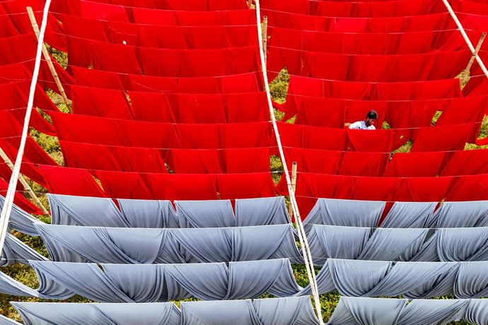 Um trabalhador pendura centenas de tecidos de cores vivas para secar ao sol após o processo de coloração. Depois de secos, os tecidos são oferecidos nos mercados atacadistas e, posteriormente, enviados para as fábricas de roupas. Cada fileira de fios é av
