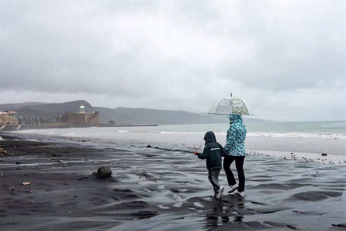 Archivo - Dos personas se cubren de la lluvia con un paraguas mientras caminan por una playa en Las Palmas de Gran Canaria, Canarias (España). 