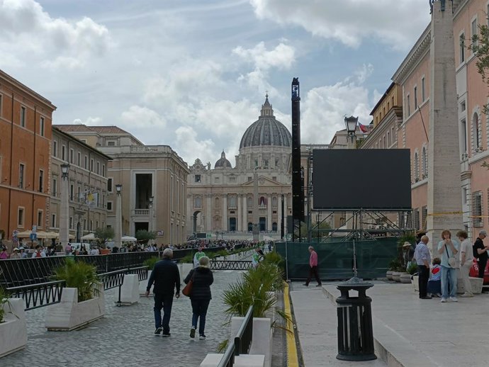 Dos personas caminan por Via de la Conciliazione junto a una pantalla gigante, de camino a la Plaza de San Pedro.