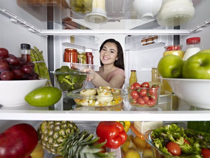 Archivo - Looking from inside a fridge full of healthy food to a young woman smiling to the camera