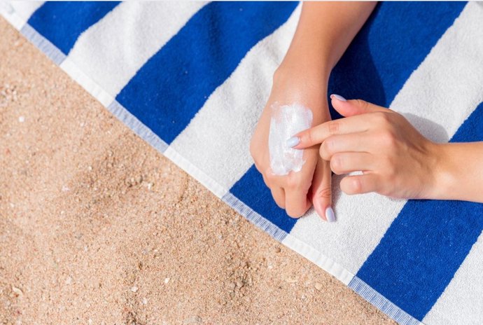 Foto recurso donde se aprecian dos manos de una persona echándose crema solar en una toalla de rayas azules y blanca.