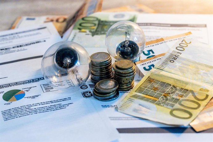 Archivo - Dos bombillas sobre billetes, monedas y facturas de la luz.