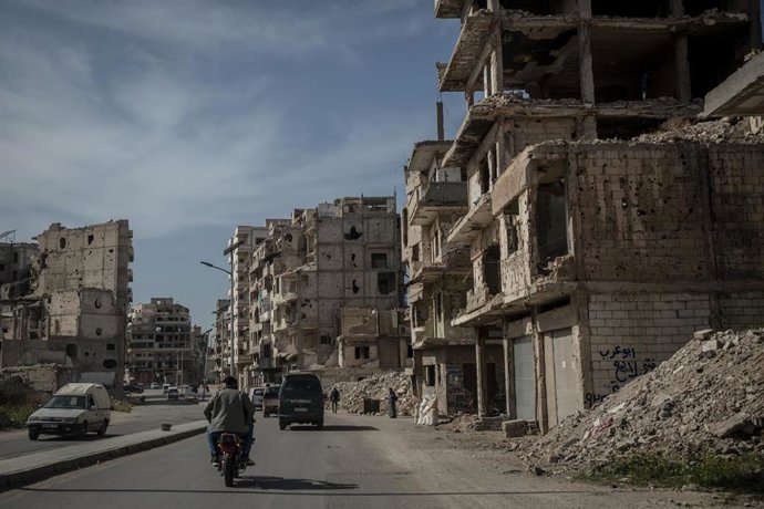 Archivo - Varios vehículos circulan por una calle con edificios dañados en Homs, Siria