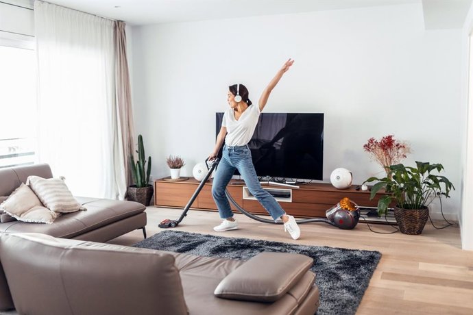 Archivo - Mujer joven y feliz escuchando y bailando música mientras limpia el piso de la sala con un limpiador de vacío