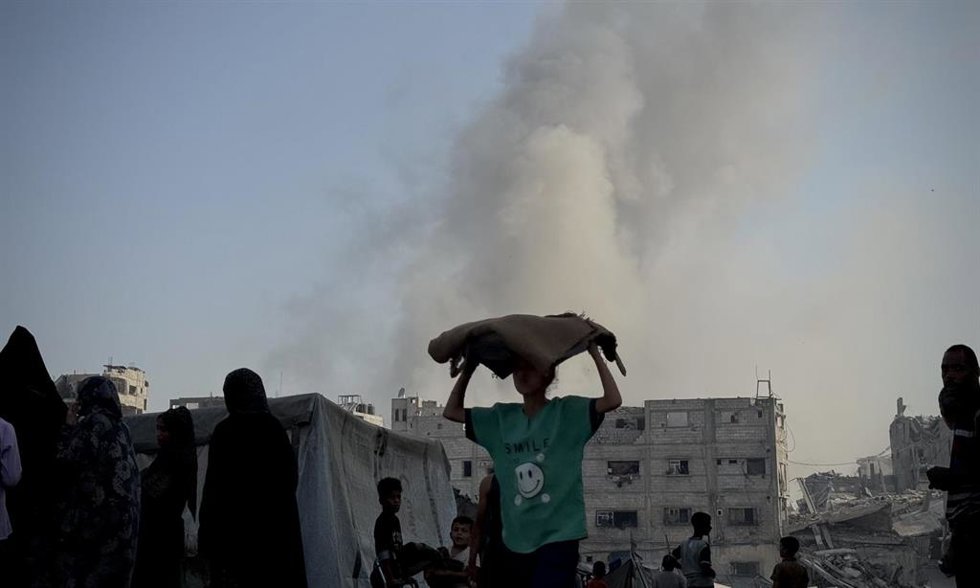 En el centro, un niño transporta un saco de comida. Al fondo, columna de humo por ataques de Israel sobre Jan Yunis, en el sur de la Franja de Gaza