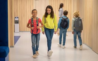 Archivo - School day. Joyful girls with backpacks walking and boys behind with their backs to camera in illuminated corridor of school