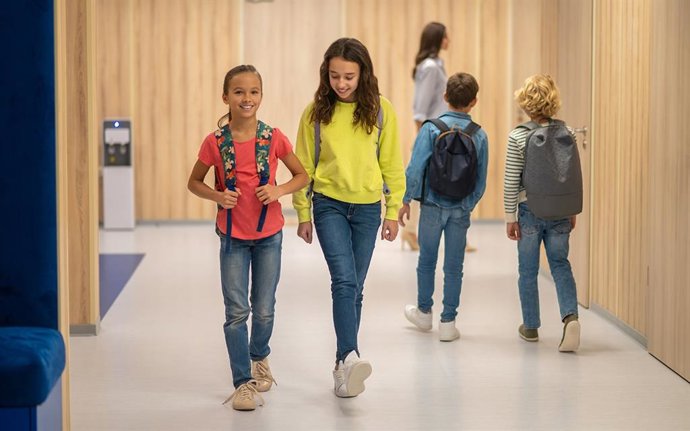 Archivo - School day. Joyful girls with backpacks walking and boys behind with their backs to camera in illuminated corridor of school