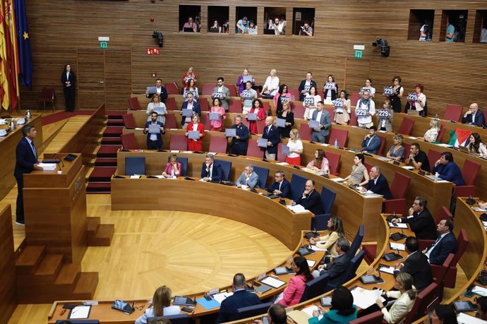 Mazón en el atril para empezar su intervención en el pleno del Debate de Política General mientras Compromís y PSPV exhiben carteles con los nombres de las víctimas mortales de la dana y la cifra