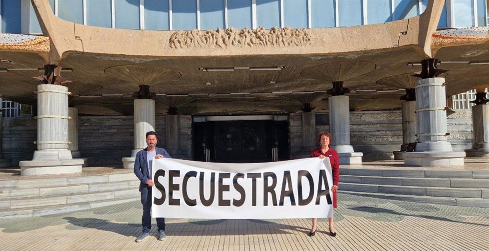 La diputada regional de Podemos-IU-AV, María Marín, y el secretario de Comunicación de Podemos, Víctor Egío, sujetan un cartel frente a la fachada de la Asamblea para denunciar la parálisis de la Asamblea