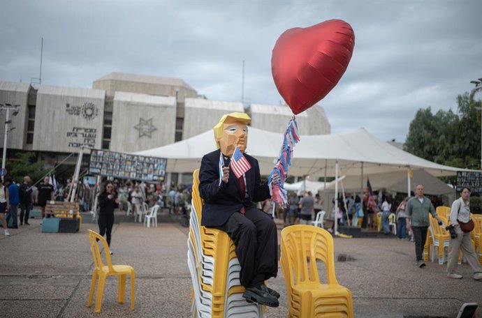 09 de outubro de 2025, Israel, Tel Aviv: Uma pessoa usa uma máscara que se assemelha ao presidente dos EUA Trump e segura a bandeira dos EUA e um balão em forma de coração enquanto as pessoas se reúnem na praça dos reféns em Tel Aviv para comemorar o acor