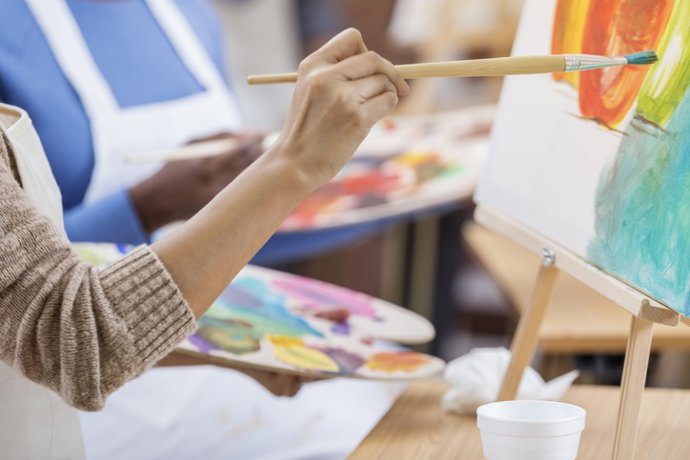 Archivo - Mujer trabaja en una pintura. Ella está usando un pincel mientras pinta sobre un lienzo. Ella sostiene una paleta. La gente está pintando de fondo.