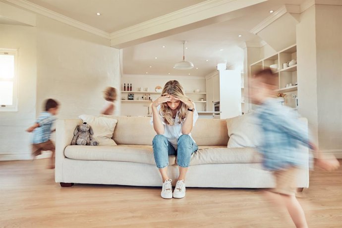 Archivo - Madre estresada sentada en el sofá mientras sus hijos corren por la sala de estar. Hijo e hija hiperactivos. Hermanos activos persiguiéndose unos a otros en la casa.  TDAH