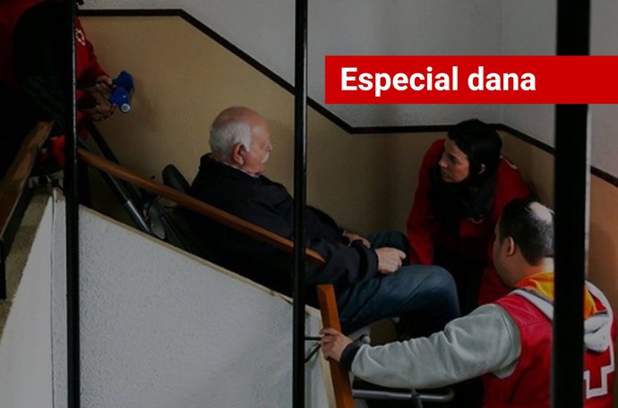 Imagen de una asistencia de Cruz Roja con silla oruga para ayudar a un hombre con movilidad reducida a salir de su vivienda, en un edificio afectado por la dana