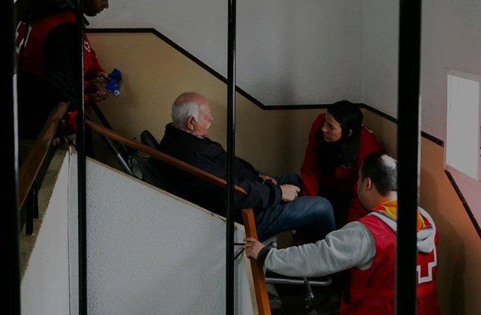 Imagen de una asistencia de Cruz Roja con silla oruga para ayudar a un hombre con movilidad reducida a salir de su vivienda, en un edificio afectado por la dana
