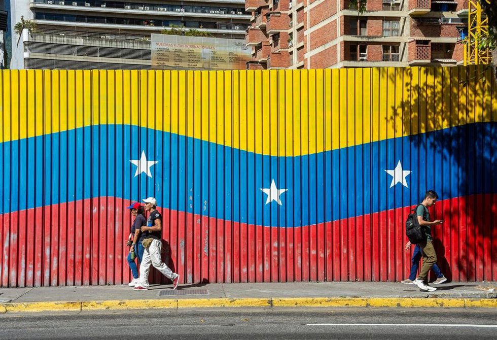 Archivo - Dos personas caminan delante de una bandera venezolana