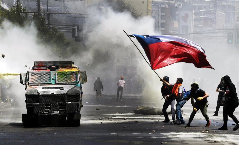 Archivo - Manifestantes odenan una bandrea chilena y lanzan piedras contra un vehículo policial que lanza gases lacrimógenos durante el estallido social de 2019 en Valparaíso (Chile)