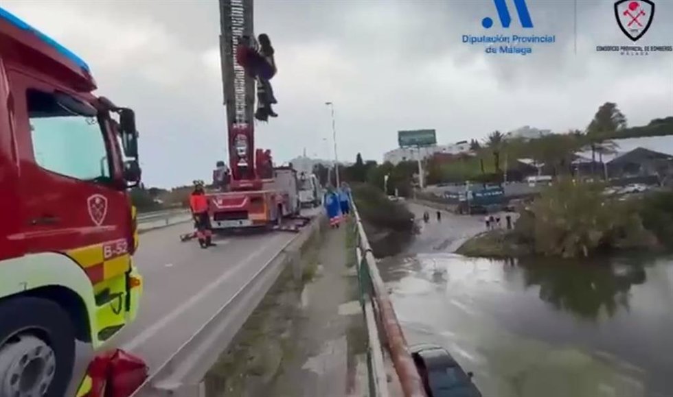 El bombero rescatador y la mujer son elevados desde el vehículo auto escala del CPB mientras el coche de la víctima se queda atrapado en el cauce del río Guadalmansa por el que la conductora pretendía avanzar a pesar de la crecida del mismo.
