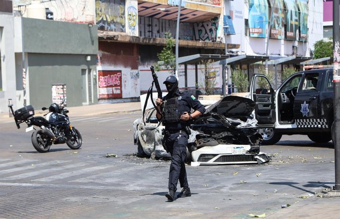 JALISCO, 23 de fevereiro de 2026 — Um policial vigia um carro queimado em uma rua de Guadalajara, capital do estado de Jalisco, no México, em 22 de fevereiro de 2026. As forças militares mexicanas mataram Nemesio Oseguera Cervantes, conhecido como “El Men