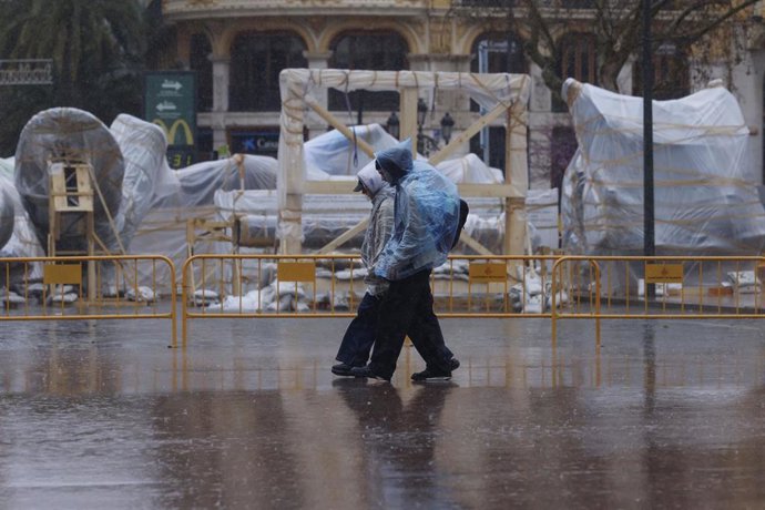 Personas caminan ante ninots tapados con plásticos por la lluvia durante el montaje de las Fallas 2026