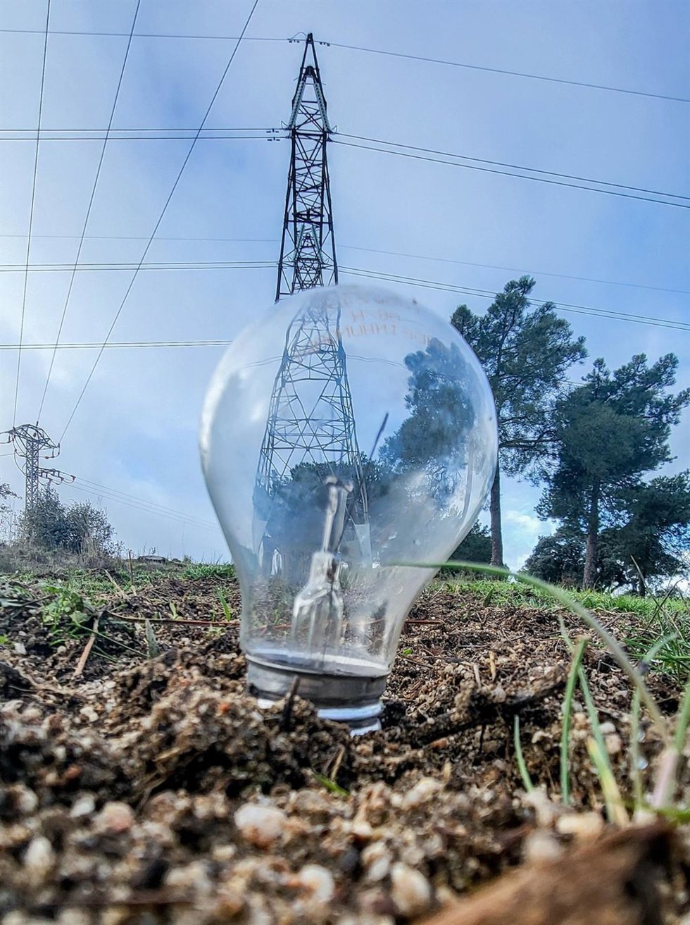 Archivo - Una bombilla clavada el césped delante de una torre de energía eléctrica