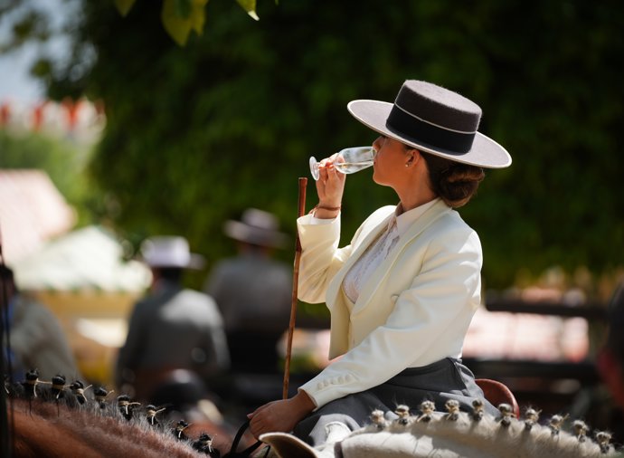 Una mujer vestida de amazona sobre un caballo, con una copa de manzanilla en la mano, por las calles del Real de la Feria, en foto de archivo.