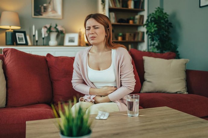 Archivo - Mujer sentada en el sofá agarrándose el estómago con dolor en el salón de casa