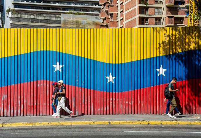 Archivo - Dos personas caminan delante de una bandera venezolana en una calle de Caracas, Venezuela