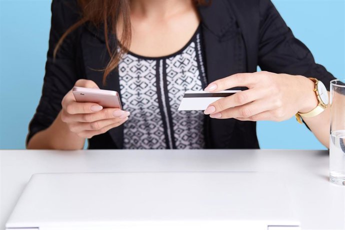 Archivo - Portrait of faceless woman wears black suit using cell phone and credit card while sitting at white table near laptop computer and glass of water, isolated over blue background. Online payment concept