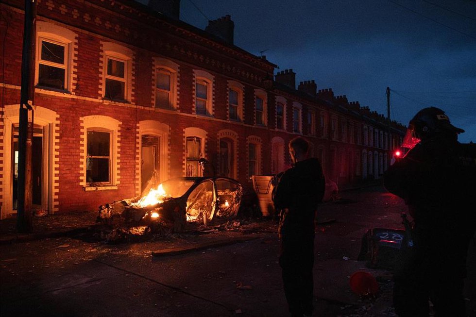 Un vehículo arde frente a unas viviendas, mientras agentes del Servicio de Policía de Irlanda del Norte (PSNI) observan desde cerca tras los disturbios ocurridos durante la noche.