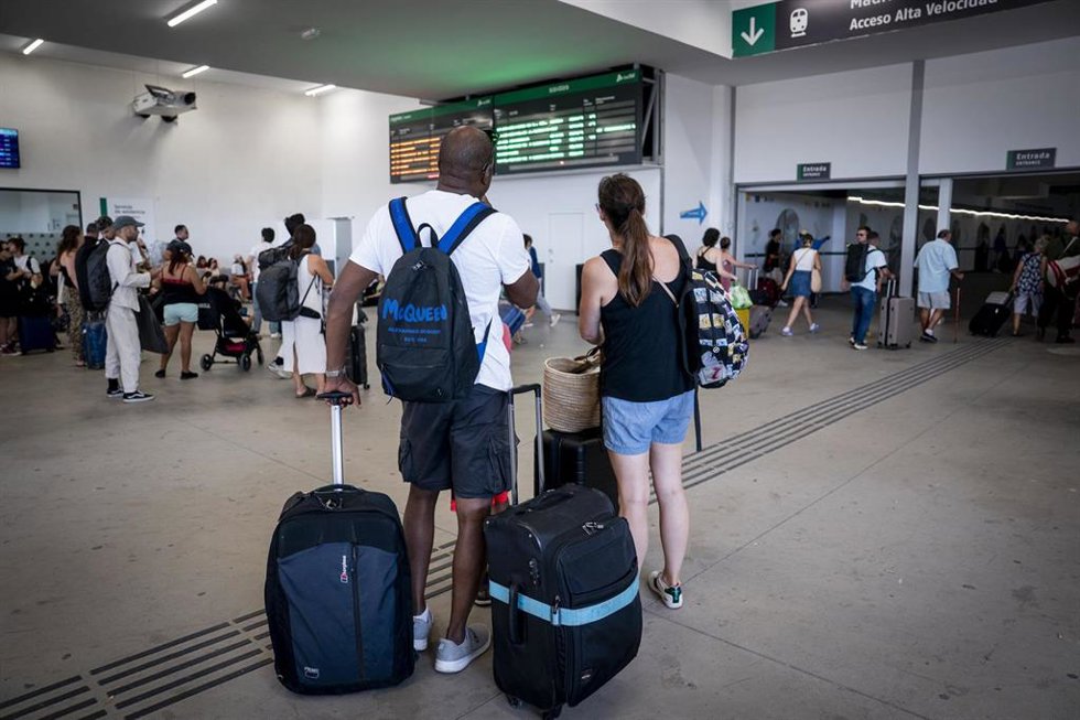 Archivo - Varias personas esperan con maletas y observan los carteles de salidas en una estación de tren