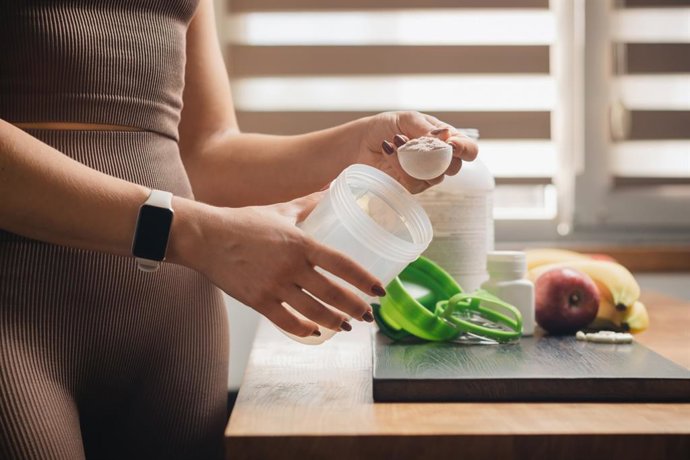 Archivo - Una mujer atlética en ropa deportiva con una cuchara medidora en la mano pone una porción de proteína de suero en polvo en una coctelera sobre una mesa de madera con cápsulas blancas de aminoácidos, plátanos y manzana. Creatina, colágeno