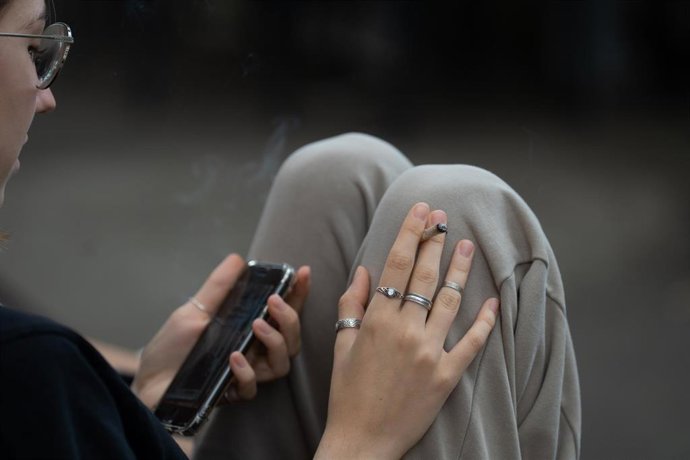 Archivo - Una joven fuma y lee su movil en Barcelona.