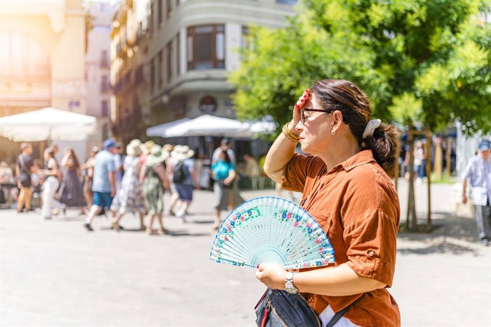 Archivo - Mujer madura caminando por la ciudad bajo el sol en un día caluroso, sufriendo un golpe de calor en un clima extremadamente caluroso, soplando aire con un ventilador de mano y tocándose la frente -