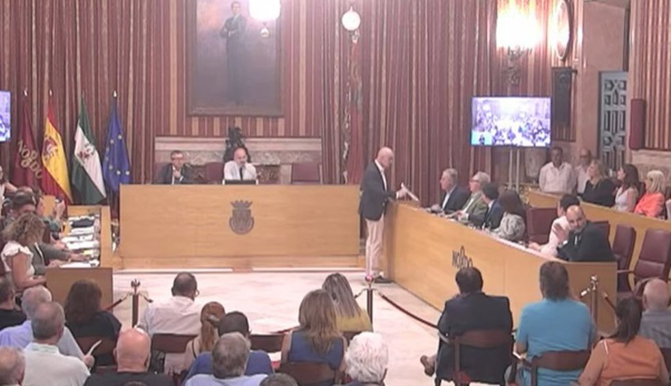 Captura de pantalla del momento del Pleno en el que Muñoz le entrega a Sanz un documento alusivo al programa municipal para menores