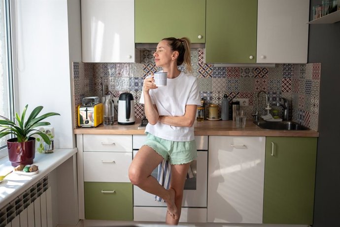 Archivo - Mujer satisfecha, feliz y relajada, de pie sola en la cocina, en un ambiente hogareño y acogedor, vestida con ropa informal, con una taza de té o café caliente. Soltera