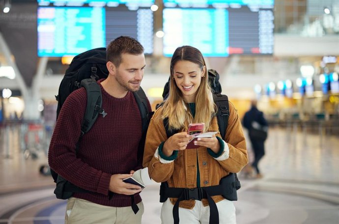 Pareja en el aeropuerto con un teléfono que muestra un billete y una aplicación de viajes; ambos lucen rostros entusiasmados, conversan y sonríen. Hombre y mujer utilizan un teléfono inteligente para reservar digitalmente hotel, taxi o autobús.